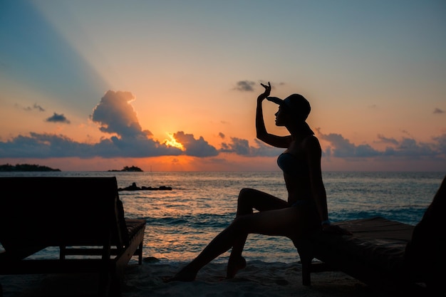Силуэт подтянутой девушки, сидящей на шезлонге на берегу океана с закатом на Мальдивах с горизонтом