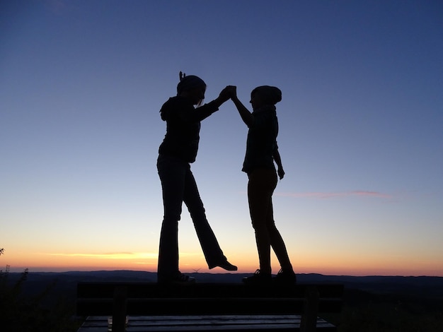 Silhouette friends dancing on bench against sky during sunset