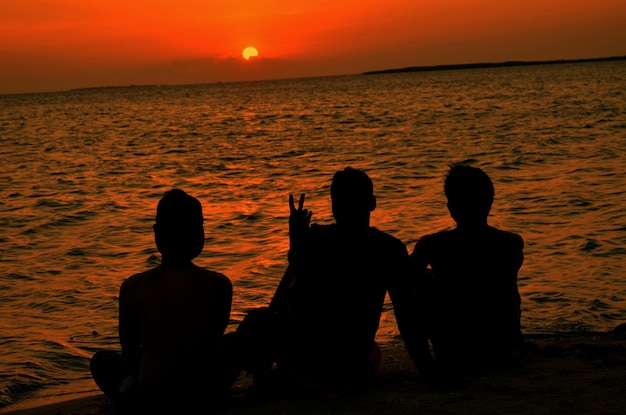 Photo silhouette friends sitting on shore at beach during sunset