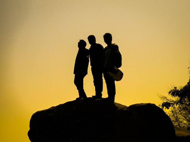 Photo silhouette friends standing on boulder against yellow sky during sunset