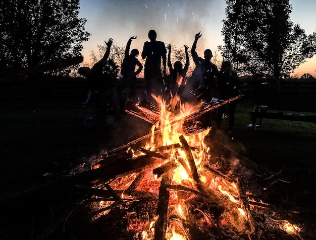 Photo silhouette friends with arms raised standing in front of campfire at sunset