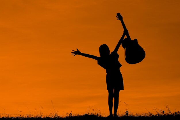 Silhouette girl holding guitar while standing on field against orange sky