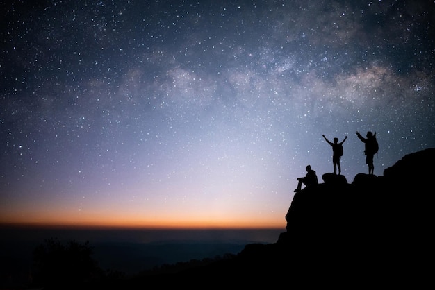 両手を広げて立っている若い旅行者のグループのシルエットで、山の頂上で星の天の川と夜空を見ました。彼らは旅行を楽しみ、頂上に到達したときに成功しました。