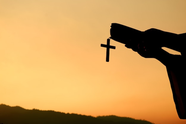 Silhouette of hand holding bible book with cross on sunset background