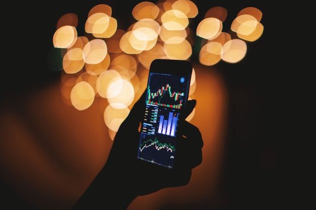 Силуэт руки, держащей смартфон с биржевой дисплеем с светлом фоне