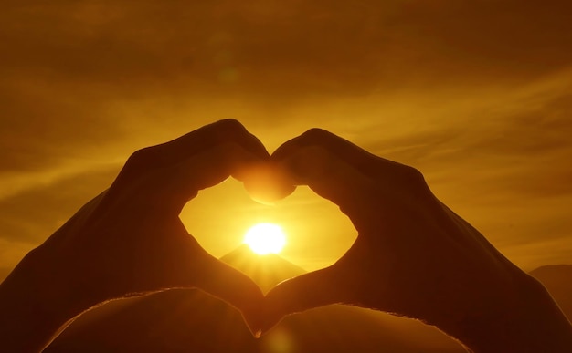 Silhouette of hand making heart sign to the bright sun rising over the Silhouette of mountain peak