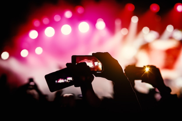 Photo silhouette of hands using smartphones to take pictures and videos at live music show.