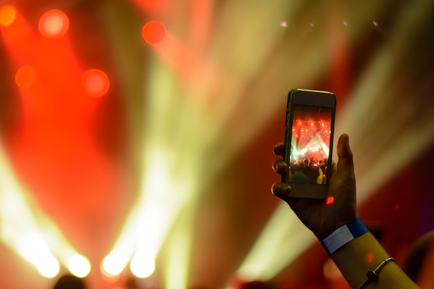 Silhouette of hands with a smartphone on the background of the singing artists in the light of the red lights