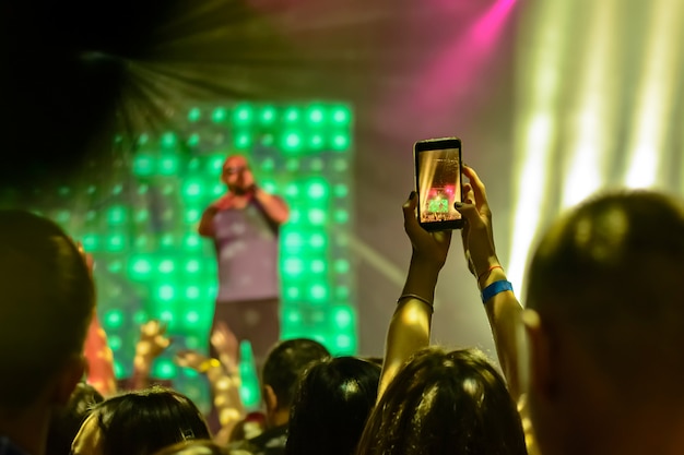 Silhouette of hands with a smartphone on the background of the singing artists in the light of the red lights