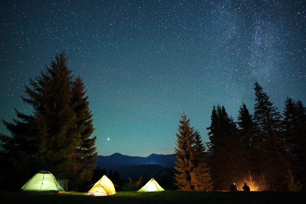 Silhouette of hikers resting besides burning bonfire near illuminated tourist tents on camping site in dark mountains under night sky with sparkling stars. Active lifestyle and outdoor living concept.