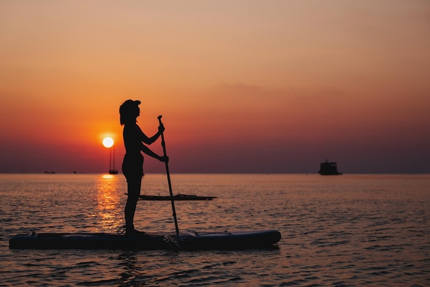 Silhouette image of a young woman on stand up paddle board in a sea before sunset