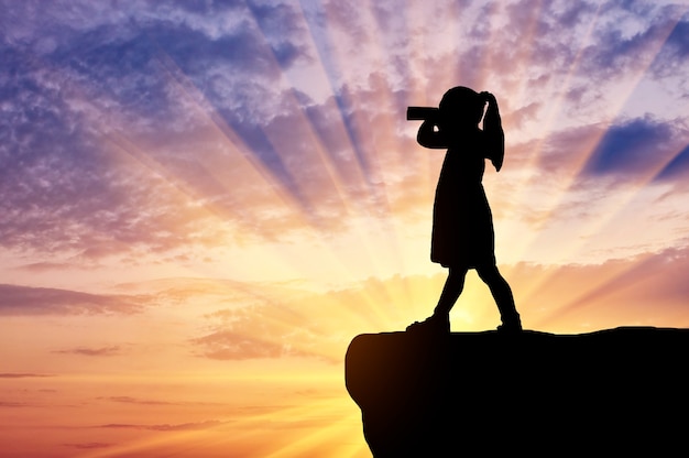 Silhouette of little girl child standing on top looking through binoculars. The concept of children's curiosity and discovery