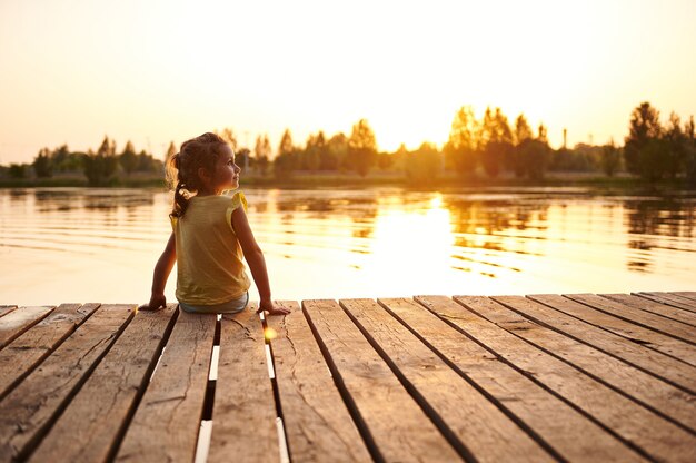 桟橋に座って、湖のほとりに夕日を見ている少女のシルエット。夕日の美しい光が湖の水に映っています。