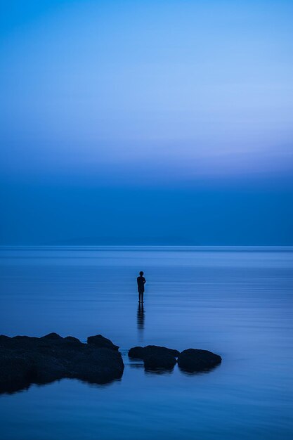 Силуэт одинокой фигуры, стоящей на берегу моря в сумерках, голубые тона сумерек.