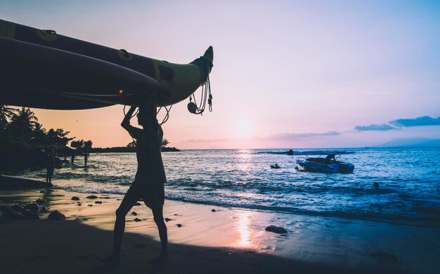 사진 해가 지는 동안 하늘을 배경으로 해변에서 배를 운반하는 실루 남자