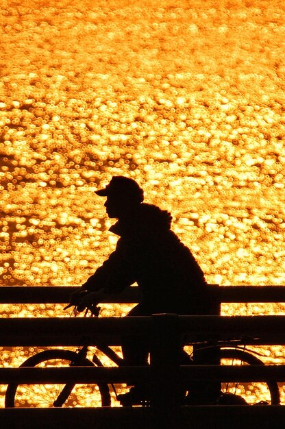 Silhouette man cycling on bridge over sea during sunset