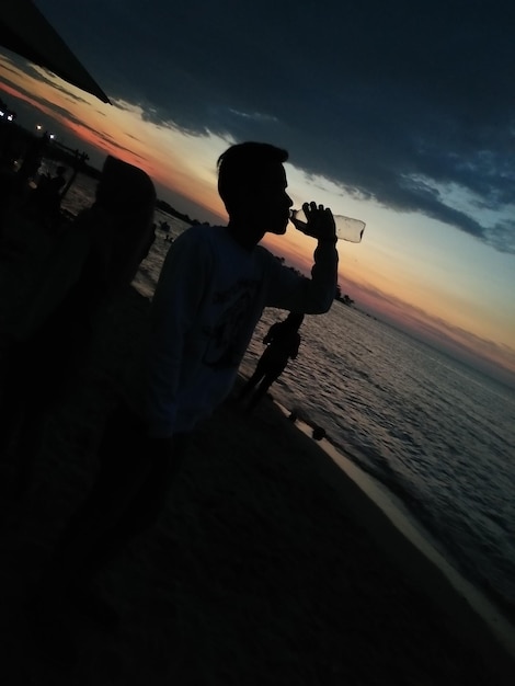 Foto uomo a silhouette che beve acqua da bottiglia sulla spiaggia