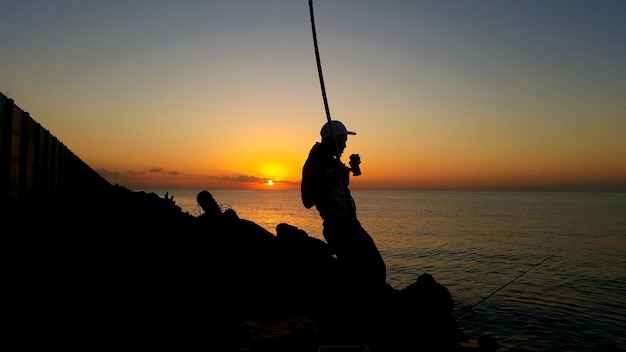 写真 夕暮れの空に向かって海で釣りをしている男性のシルエット