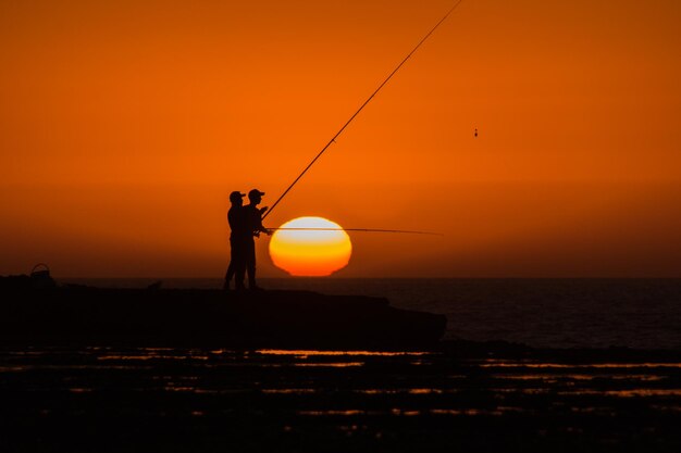 Silhouette man fishing by sea against orange sky