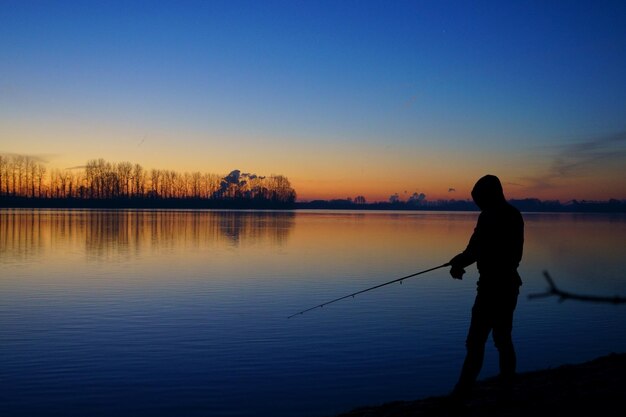 写真 夕暮れの空を背景に湖で釣りをしている男性のシルエット