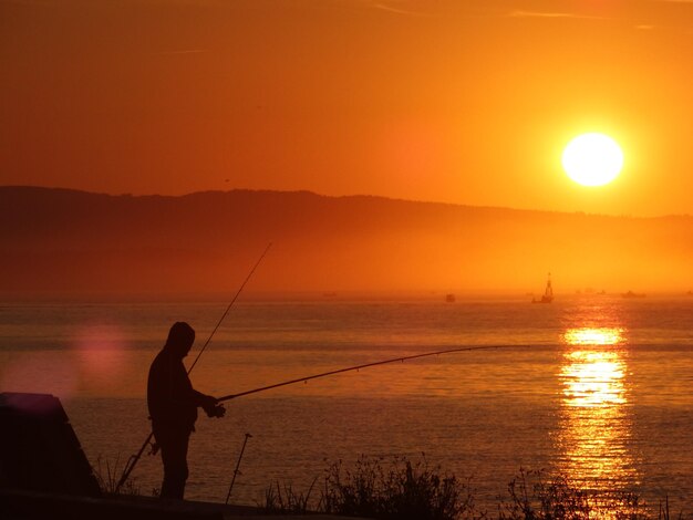 Silhouette man fishing in sea against orange sunset sky