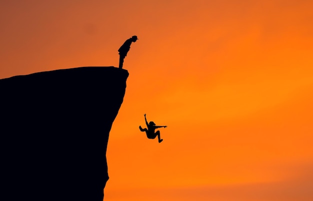 Silhouette man looking at woman falling from cliff against clear orange sky