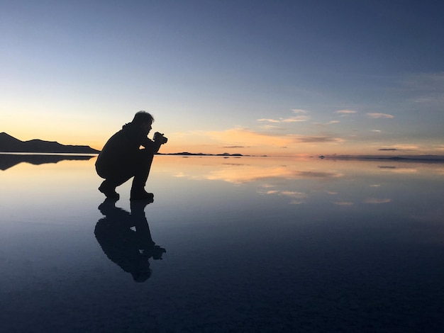 Силуэт мужчины фотографирует на пляже на фоне неба во время захода солнца