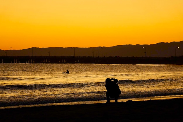 Silhouette man photographing friend at beach against clear sky during sunset