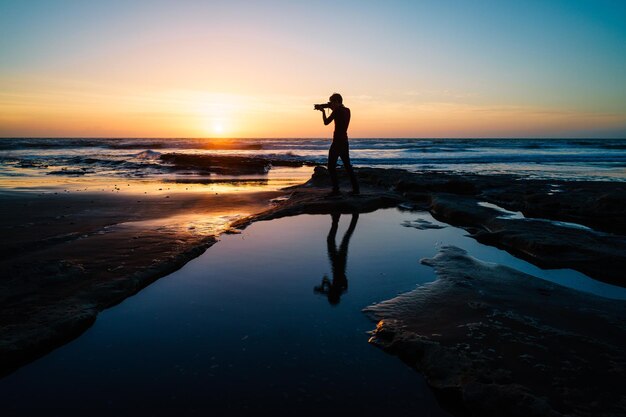 Photo silhouette man photographing while standing at beach against sky during sunset