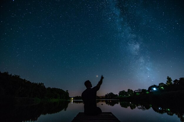 写真 夜の空を背景に湖岸を指しているシルエットの男