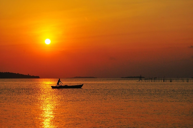 Silhouette man sailing boat in sea against orange sky during sunset