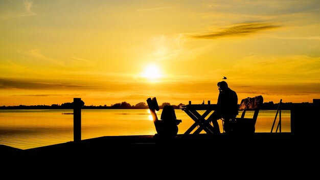 Silhouette man sitting by lake against orange sky