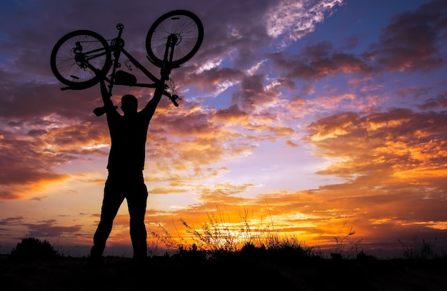 Silhouette the man stand in action lifting bicycle above his head at sunset