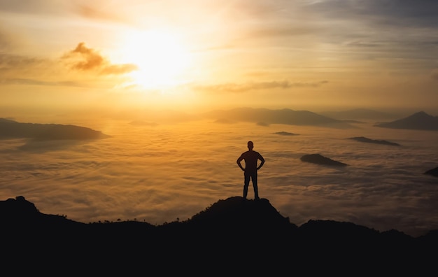 Silhouette man standing and akimbo on peak of stone rock in\
mountain and watching to sunrise fog mist sunset and sunlight misty\
valley man hikeperson silhouette stand mountain nature concept