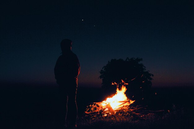Silhouette man standing by bonfire on field against sky at night