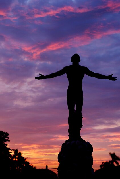 Silhouette man standing by statue against sky during sunset