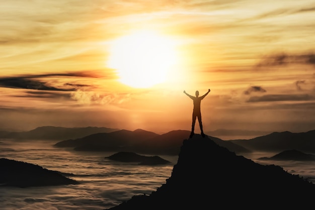 Silhouette man standing and raise two hand on peak stone rock\
in mountain and watching to sunrise fog mist sunset sunlight misty\
valley man hikeperson silhouette stand mountain nature concept