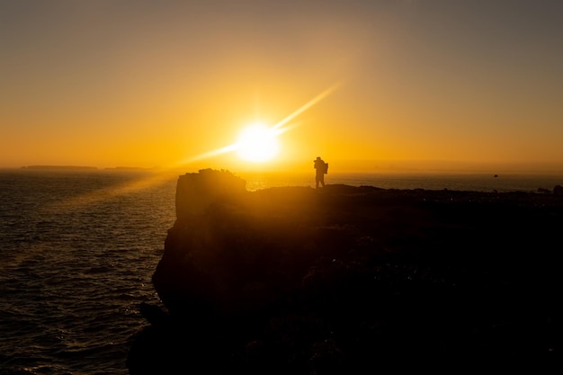 Силуэт человека, стоящего на вершине скалы и фотографирующего море на закате
