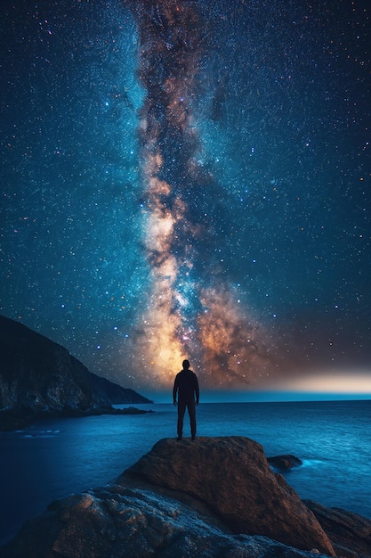 silhouette of man standing on top of a mountain on background of a starry night sky with bright Milky way and sea water