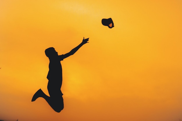 Silhouette man throwing cap while jumping against sky at sunset