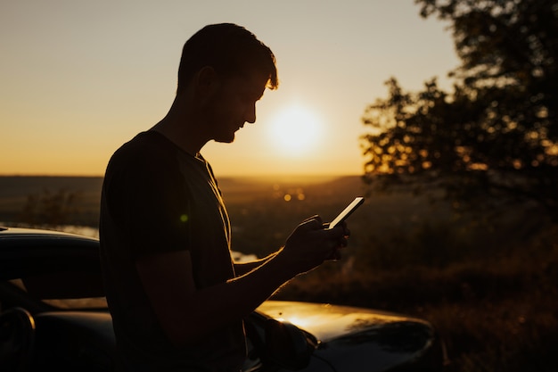 車で旅行し、壮大な夕日の丘の上に携帯電話で立っている男のシルエット。