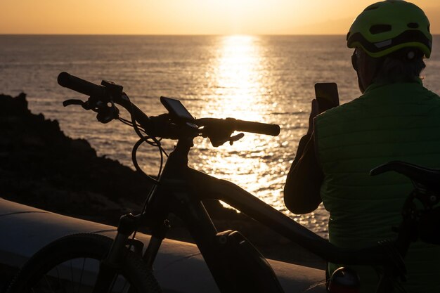 Silhouette of man using mobile phone at sunset wearing helmet after cycling along the sea