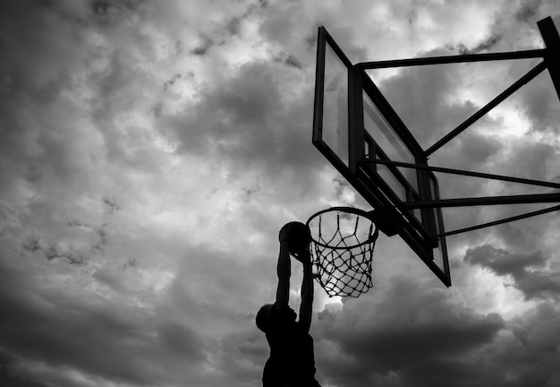 Silhouette di un uomo che lancia una palla in un canestro da basket per strada contro un cielo con nuvole di colore bianco e nero