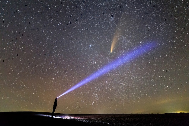 Silhouette of a man with flashlight on his head pointing bright beam of light on starry sky with Neowise comet with light tail.