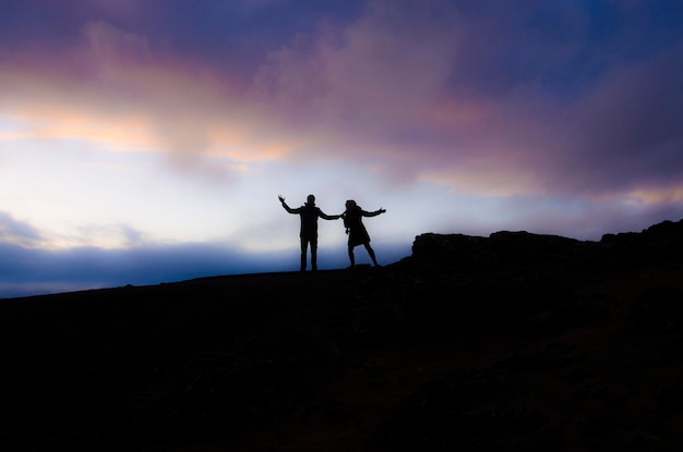 silhouette of man and woman with arms outstretched while standing on landscape against sky during sunset