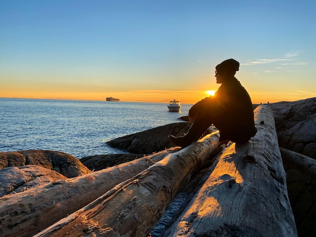 写真 夕暮れの時に木材の上に座って水を見ている男のシルエット