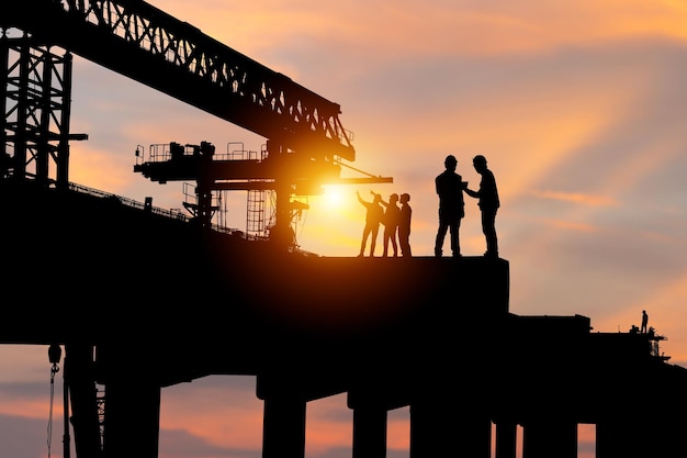 Фото Силуэт инженера и рабочего, проверяющих проект на строительной площадке фон строительной площадки при заходе солнца вечером