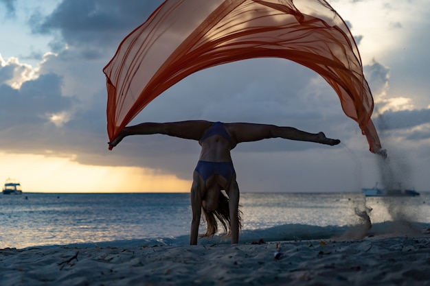 Фото Силуэт гибкой подходящей женщины, делающей стойку на руках с шелком во время драматического заката с грозовыми облаками на фоне морского пейзажа, концепция счастья, свободы и беззаботности