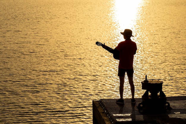 写真 夕日の下で川のギターを弾くギタリストのシルエット