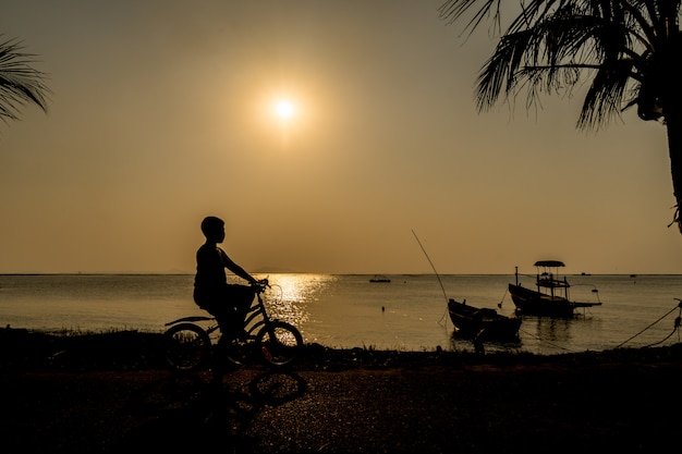 写真 彼の休日休暇のリラックスできる時間のビーチの近くの夕日の下で自転車に乗る少年のシルエット